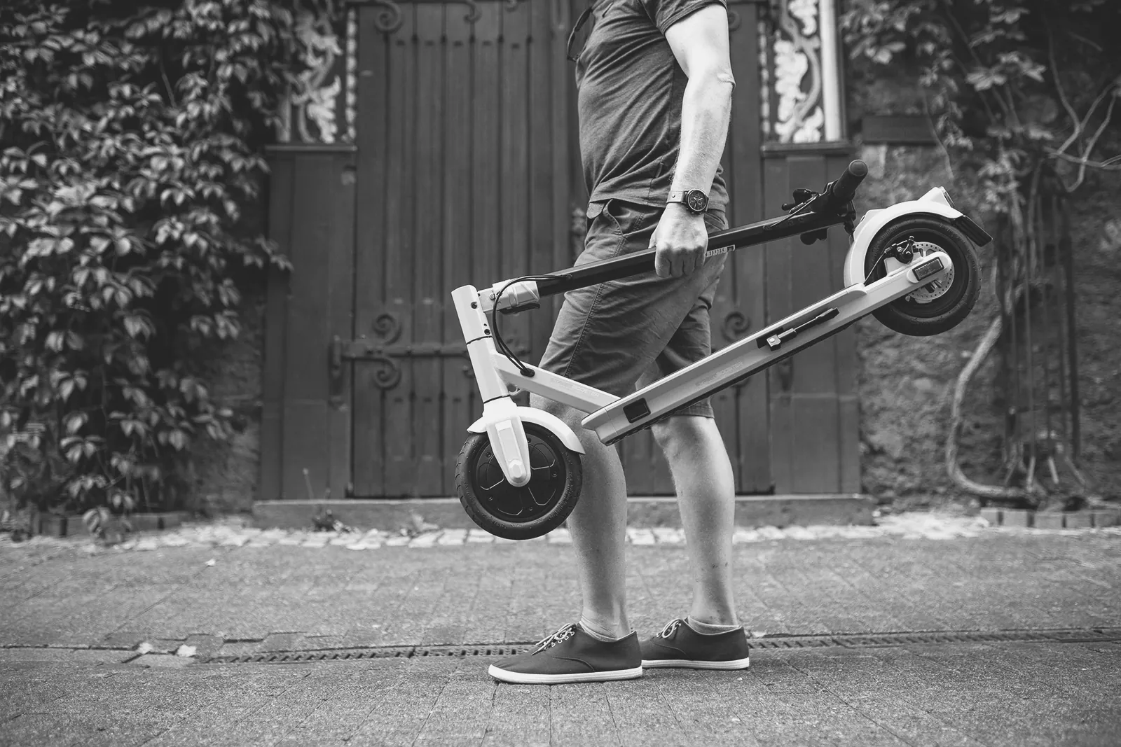 Mann trägt einen zusammengeklappten E-Scooter über das Kopfsteinpflaster